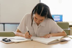 A dark-haired female student reads a book and takes notes seated at a library table. 